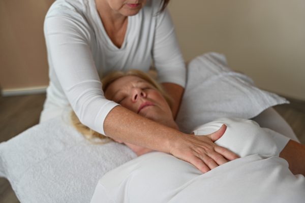 Osteopathie. Die Patientin liegt auf der Behandlungsliege auf dem Rücken. Die Osteopatin sitzt hinter ihrem Kopf und hat eine Hand auf den Brustkorb und eine Hand unter den Nacken der Patientin liegen.